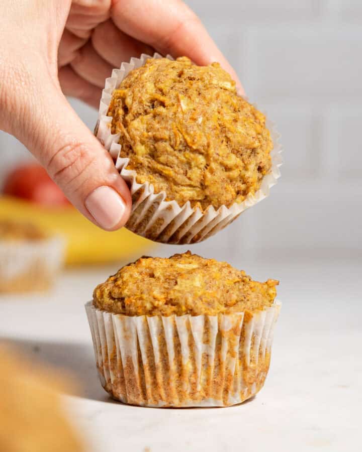 Two apple banana carrot muffins on a table with one muffin being held above the other to show the soft texture.