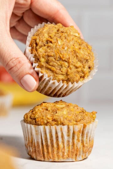 Two apple banana carrot muffins on a table with one muffin being held above the other to show the soft texture.