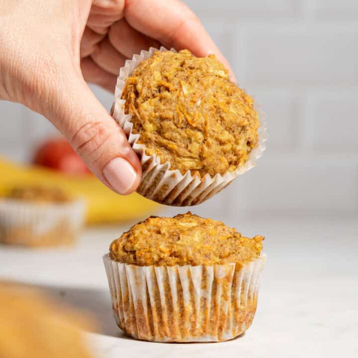 Two apple banana carrot muffins on a table with one muffin being held above the other to show the soft texture.