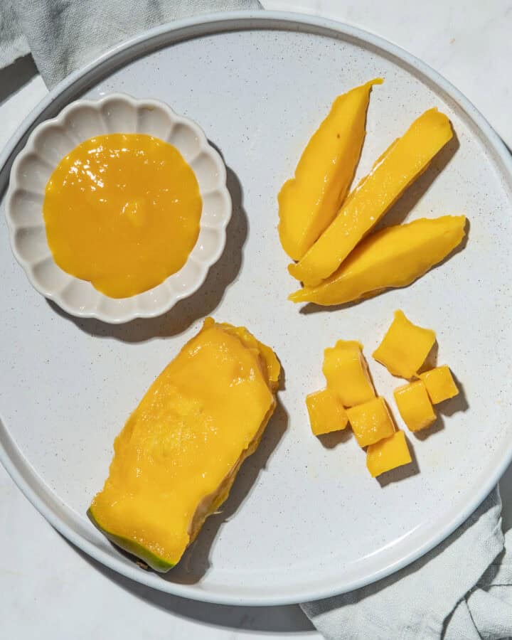 Mango pit with most of the flesh removed, mango spears and bite-sized mango pieces and a small bowl of mango puree on a plate.