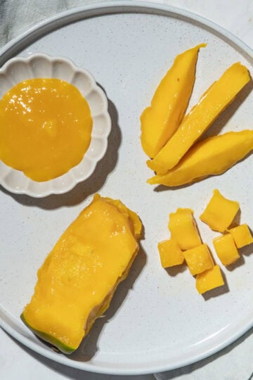 Mango pit with most of the flesh removed, mango spears and bite-sized mango pieces and a small bowl of mango puree on a plate.