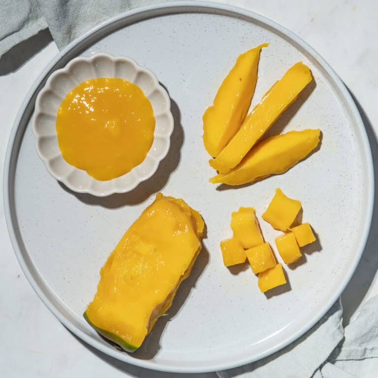 Mango pit with most of the flesh removed, mango spears and bite-sized mango pieces and a small bowl of mango puree on a plate.