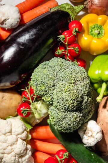 A lot of veggies on a marble background. Among them: broccoli, bell peppers, aubergine, potatoes, sweet potatoes, tomatoes, onion, garlic, carrots, cauliflower and zucchini.
