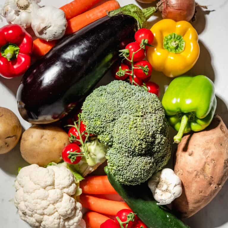 A lot of veggies on a marble background. Among them: broccoli, bell peppers, aubergine, potatoes, sweet potatoes, tomatoes, onion, garlic, carrots, cauliflower and zucchini.