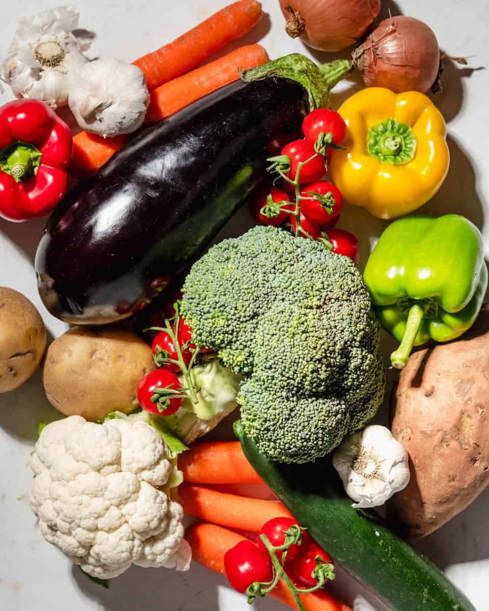 A lot of veggies on a marble background. Among them: broccoli, bell peppers, aubergine, potatoes, sweet potatoes, tomatoes, onion, garlic, carrots, cauliflower and zucchini.