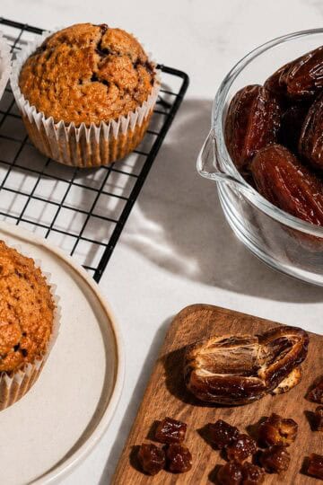 Fresh Medjool dates in a glass bowl alongside chopped dates and homemade date muffins on a cooling rack and plate, styled on a white marble surface with bright natural light and hard shadows.
