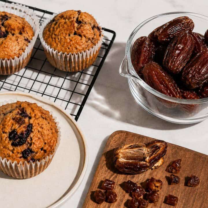 Fresh Medjool dates in a glass bowl alongside chopped dates and homemade date muffins on a cooling rack and plate, styled on a white marble surface with bright natural light and hard shadows.