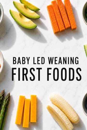 Flat lay of baby-led weaning first foods arranged on a white marble background, including broccoli, avocado, carrot sticks, sweet potato sticks, peas, cucumber spears, asparagus, cheese sticks, blueberries, and banana. Bold black text in the center reads "Baby Led Weaning First Foods," with bright natural light casting soft shadows from the right.